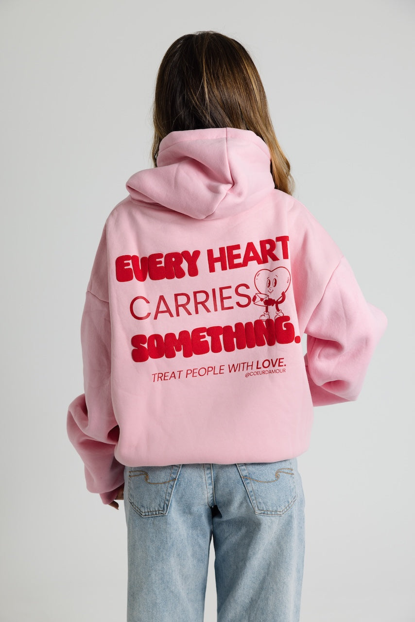 Frau von hinten, trägt ein rosa Oversize Hoodie mit rotem Backprint "Every Heart carries something". Dazu eine hellblaue Jeans vor neutralem Hintergrund.