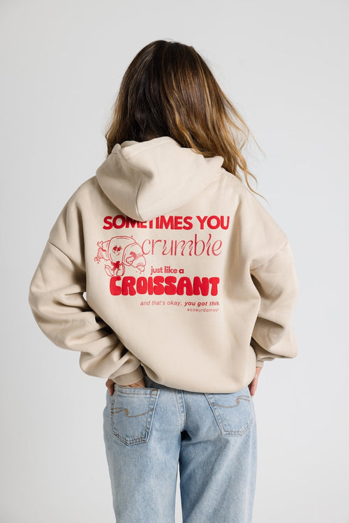 Beiger Oversize-Hoodie mit rotem Aufdruck und Croissant-Grafik, getragen von einer Frau mit offenen Haaren, Hände in den Hosentaschen