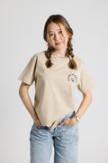 Frau mit Zöpfen trägt ein beiges Oversize T-Shirt mit schwarzem "Coeur d'Amour" Logo auf der linken Brust. Dazu eine hellblaue Jeans vor neutralem Hintergrund